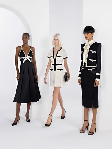 Trois modèles féminins posant dans un studio blanc avec des arches, portant des tenues élégantes en noir et blanc avec des accents blancs contrastants, la tenue centrale étant un ensemble de style safari