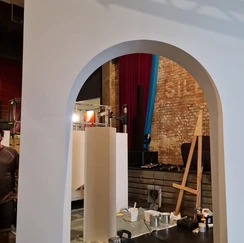 Vue à travers une arche blanche sur un atelier d'art avec un mur de briques apparentes, des rideaux colorés, et divers outils et matériaux artistiques éparpillés