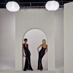 Deux modèles féminins en robes de soirée noires élégantes, posant de dos l'une à l'autre sous un arc blanc dans un studio éclairé par des lampes sphériques au plafond