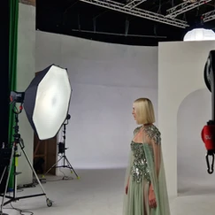 Modèle féminin en robe longue verte décorée de paillettes, posant dans un studio de photographie avec un fond blanc et un écran vert, éclairée par un grand parapluie de studio